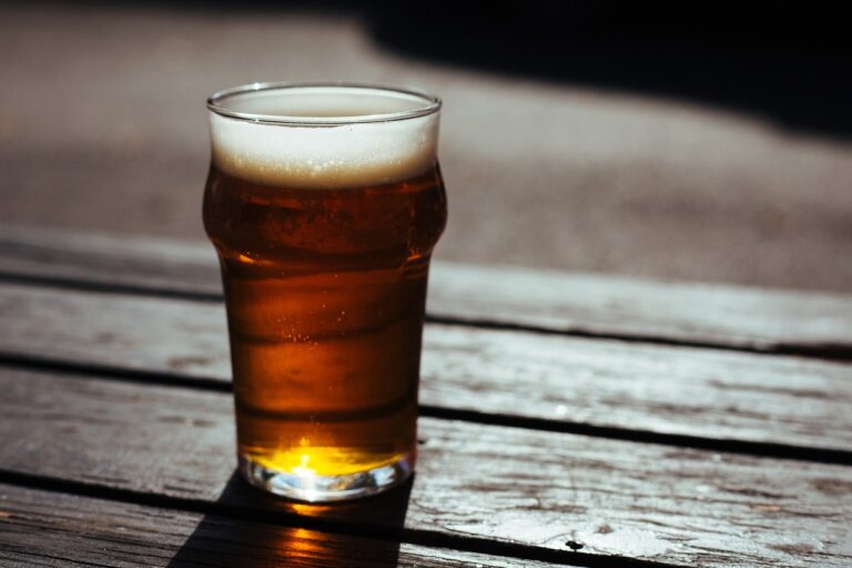 A pint glass of beer with a frothy head, placed on a wooden table with sunlight casting shadows over it.
