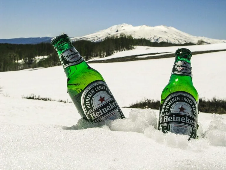 Two Heineken beer bottles are chilling in the snow with a snow-covered mountain in the background under a clear blue sky.