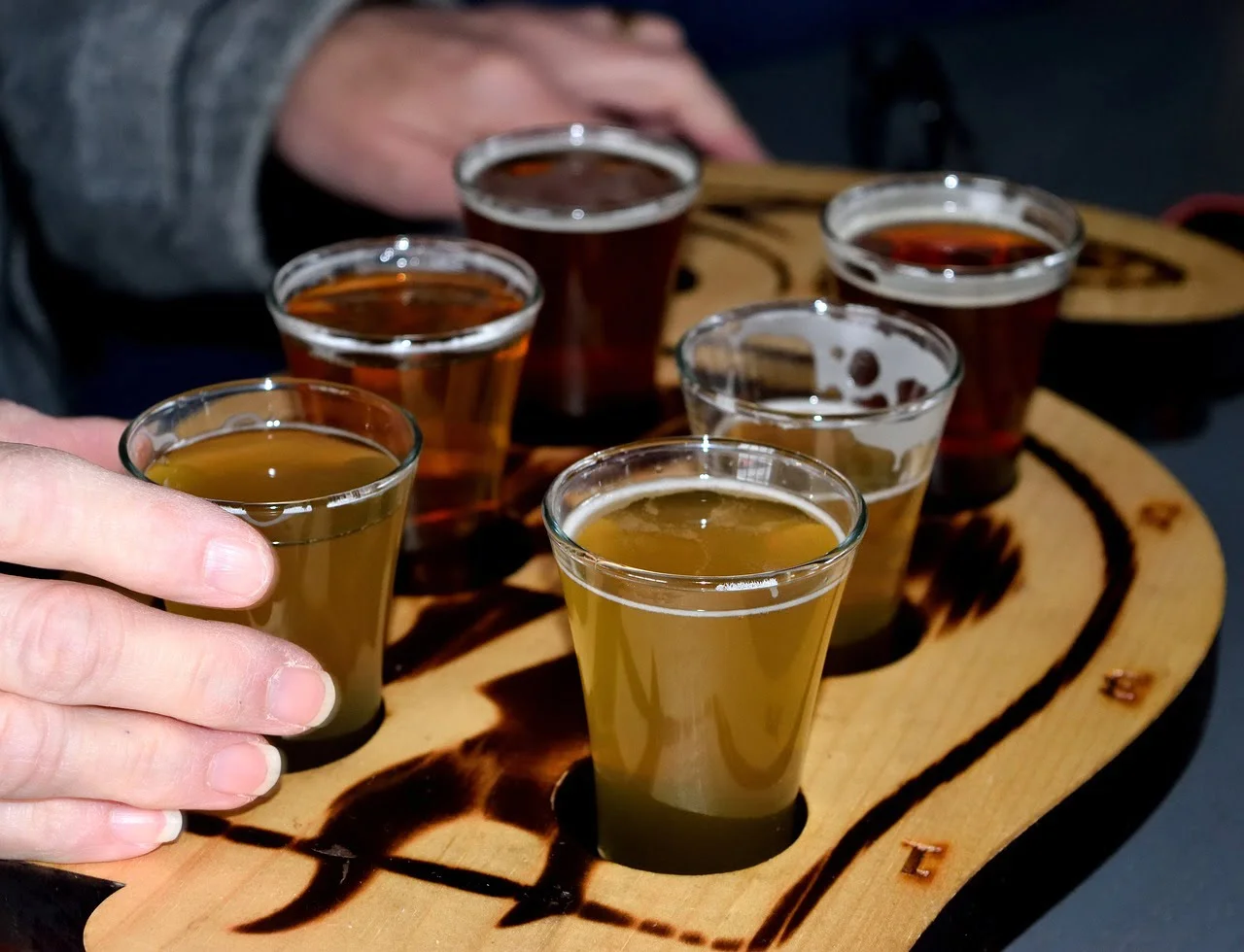 A flight of various beers in clear glasses, arranged on a wooden paddle with a hand reaching for one glass.