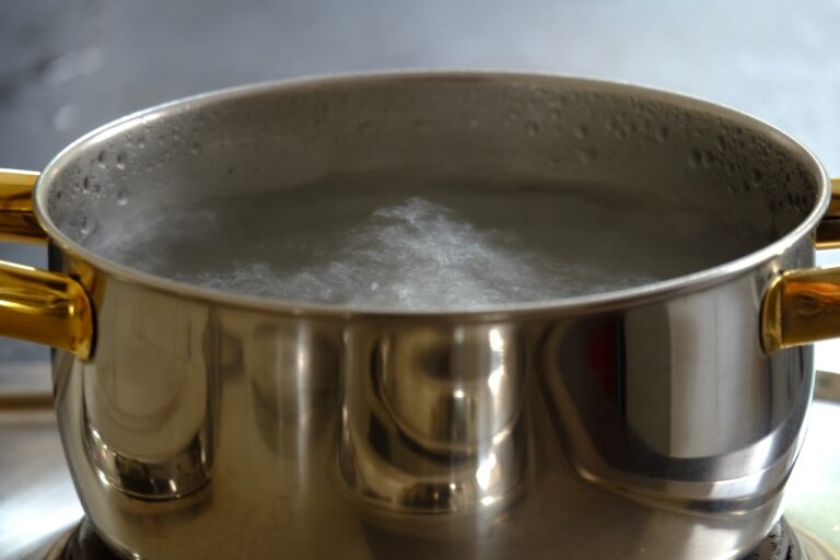 A stainless steel pot with water boiling on a stovetop, steam rising from the surface, and condensation on the inside of the pot.