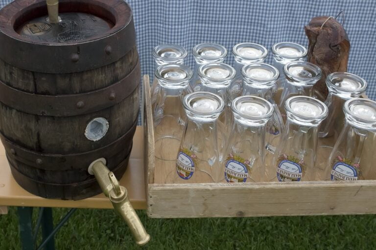A wooden barrel with a tap sitting on a table next to a tray of upside-down glass beer mugs.