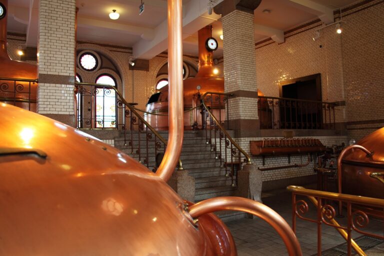 Interior of a brewery with copper brewing kettles, a staircase leading to an upper floor, tiled walls, and stained glass windows.