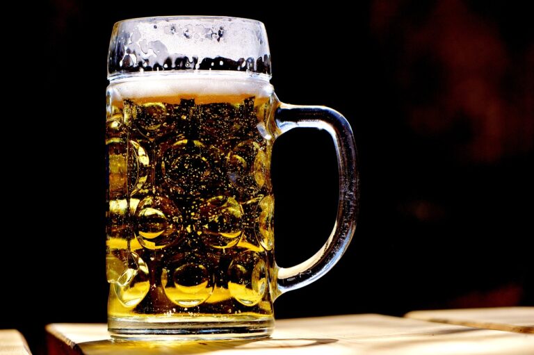 A close-up of a full beer mug with a frothy head on a wooden surface, backlit with a dark background.
