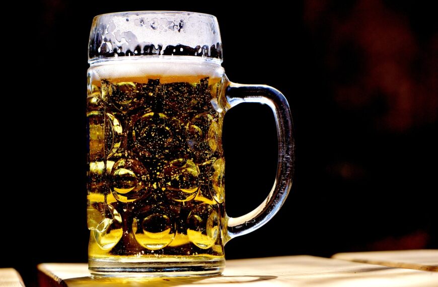 A close-up of a full beer mug with a frothy head on a wooden surface, backlit with a dark background.