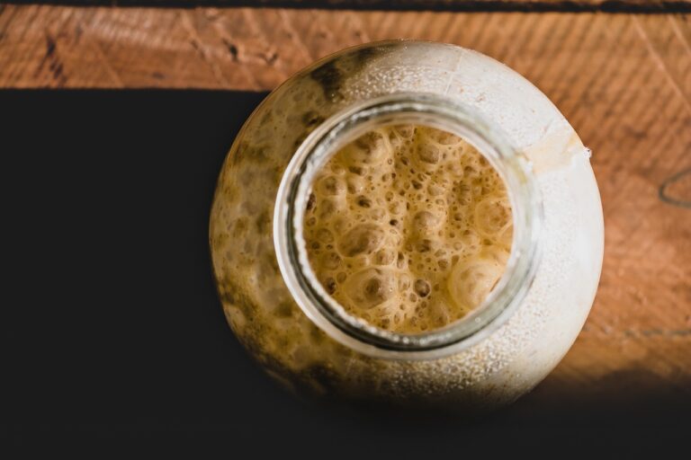 A close-up of a frothy beverage in a glass jar on a wooden surface, with the foamy texture of the drink prominently visible from the top view.