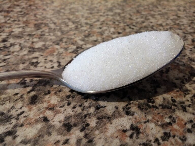 A heaping spoonful of white granulated sugar on a stainless steel spoon, set against a mottled beige countertop.