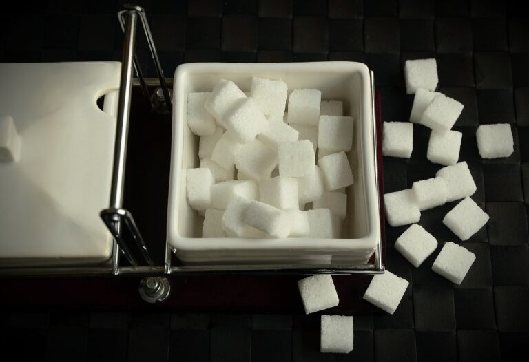 A sugar dispenser filled with white sugar cubes on a black tiled surface, with additional cubes scattered beside it.
