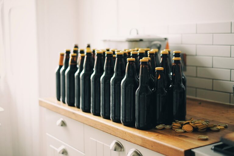 A row of brown glass beer bottles with yellow caps on a wooden countertop, with some bottle caps lying around, against a white subway-tiled kitchen backsplash.