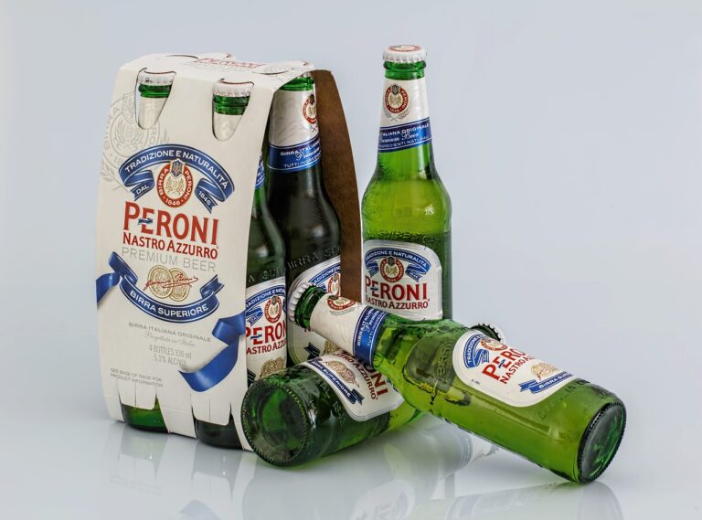 Six-pack of Peroni Nastro Azzurro beer with four bottles standing upright and one lying on its side in front of the packaging on a reflective surface.