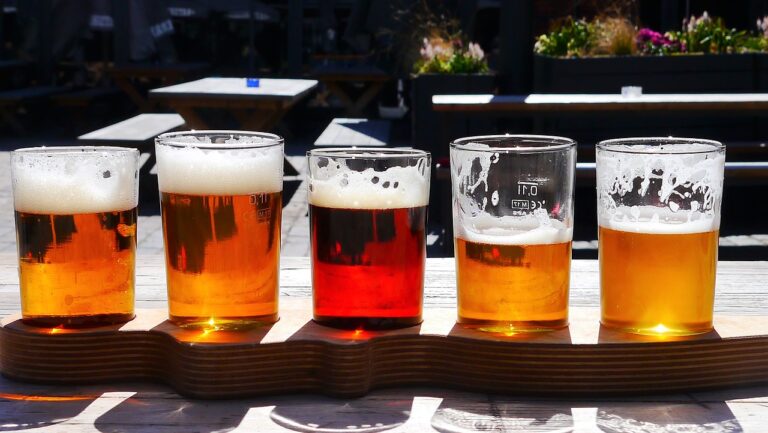 A flight of beers with varying colors and head sizes served on a wooden paddle on an outdoor table, with a sunny, blurred background.