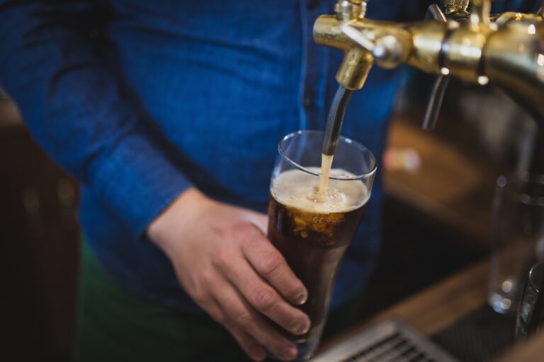 A person pours a dark carbonated beverage from a tap into a tall glass, creating a frothy head on the drink.
