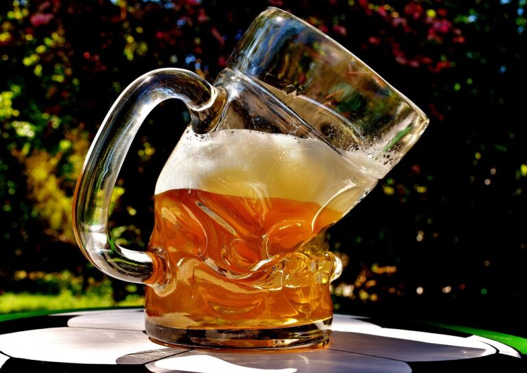 A close-up of a beer jug tipping over with beer spilling out, on an outdoor table with a blurry natural background.