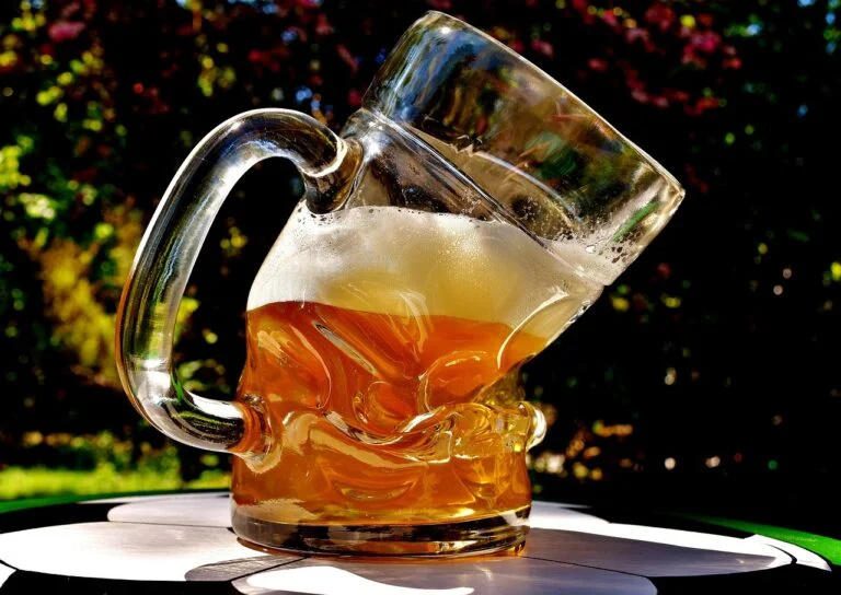 A close-up of a beer jug tipping over with beer spilling out, on an outdoor table with a blurry natural background.