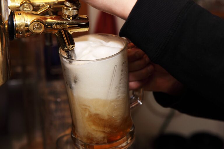 A close-up of a beer tap dispensing foamy beer into a clear mug held by a person's hand.