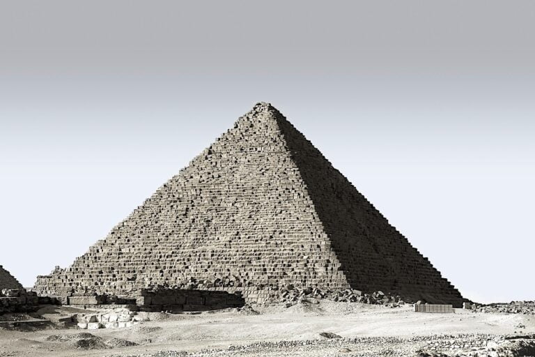 A sepia-tone photograph showing the detailed texture of an ancient Egyptian pyramid against a clear sky, with surrounding desert terrain.