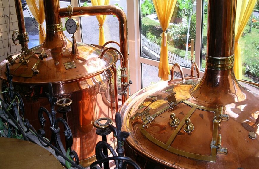Two large, polished copper brewing kettles with pressure gauges and pipes, situated in a room with sunlight streaming through windows with yellow curtains.