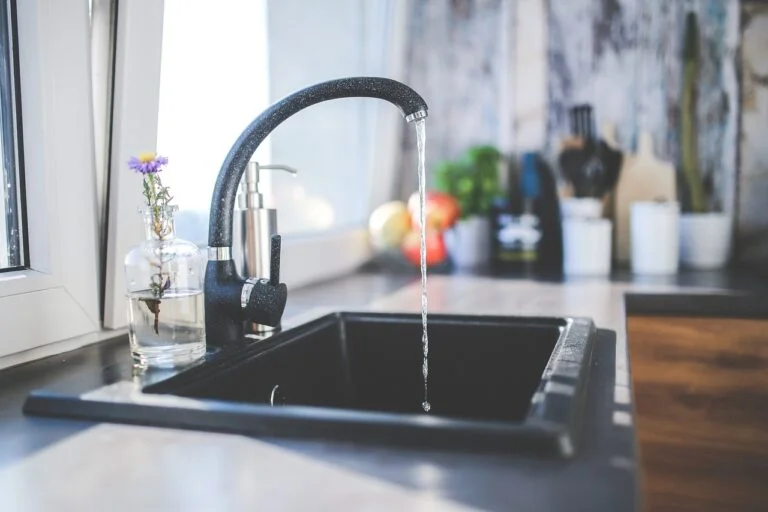 A modern kitchen with a black faucet running water into a black sink, next to a window with a clear vase holding a purple flower. The countertop is adorned with various kitchen items and potted plants.