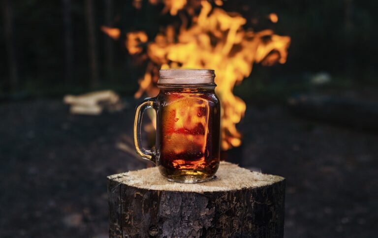 A mason jar mug filled with beer on a wooden stump with a campfire blazing in the background.