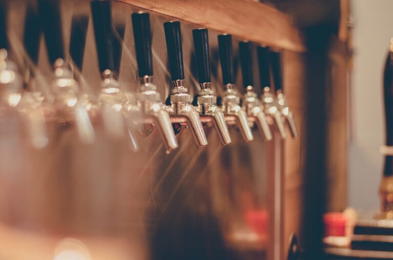 Rows of metallic beer taps with black handles mounted on a wooden structure in a blurry bar setting.