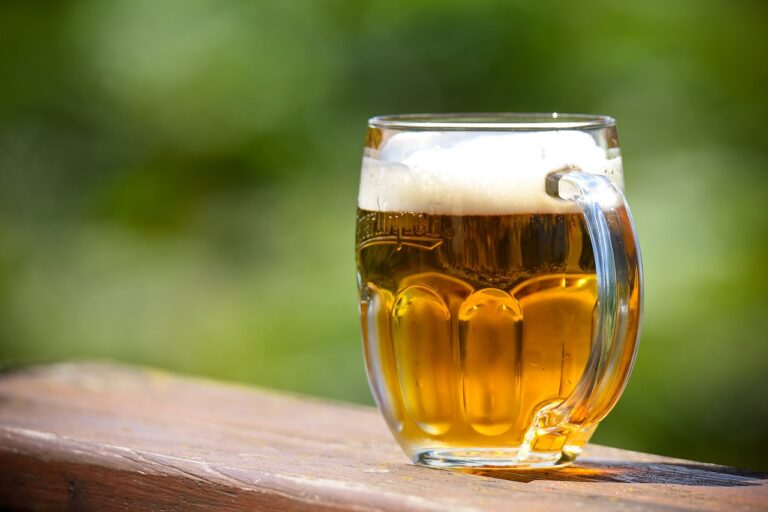 A mug of beer with foam on top, placed on a wooden surface with a blurred green background.