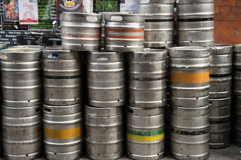 A stack of used metal beer kegs in various sizes with branding labels, placed against a wall with posters.