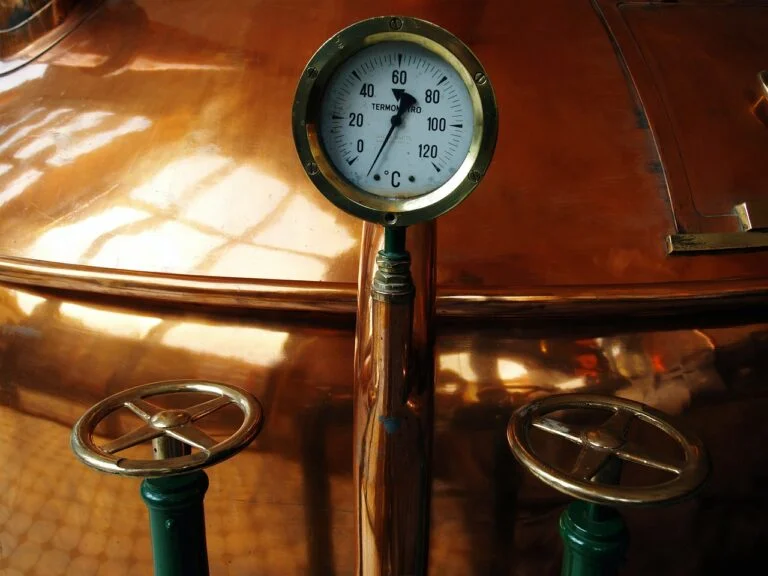 A temperature gauge reading between 20-40 degrees Celsius mounted on a shiny copper surface with two valve wheels in the foreground.