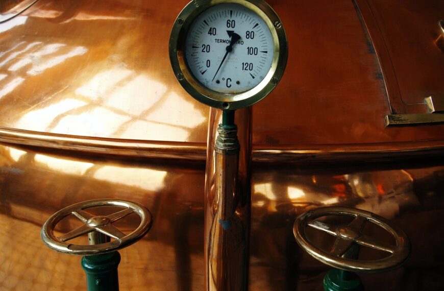 A temperature gauge reading between 20-40 degrees Celsius mounted on a shiny copper surface with two valve wheels in the foreground.