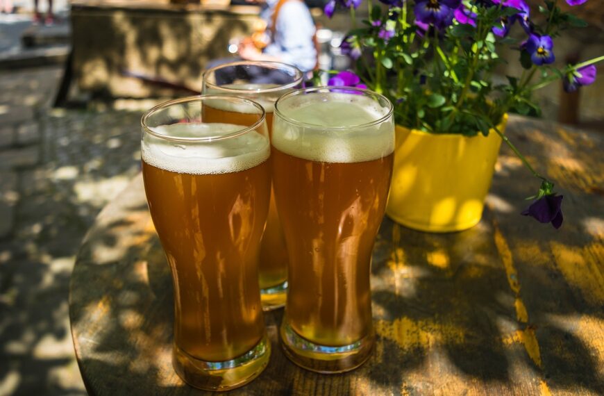 Three glasses of beer with a white frothy head on an outdoor wooden table next to a yellow pot of purple flowers, with sunlight and shadows dappling the surface and blurred background of a casual social setting.