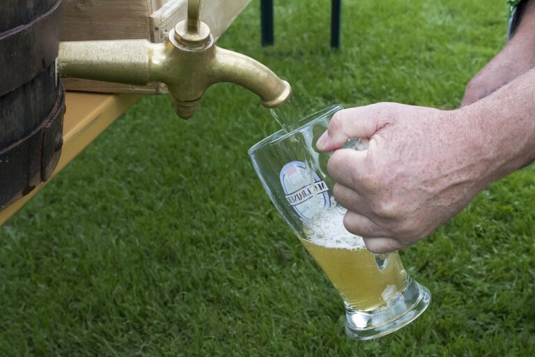 A person is pouring a golden beer from a tap on a wooden barrel into a clear glass mug with a logo, set against a background of green grass.