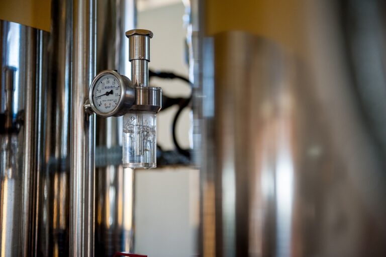 A close-up photo of a pressure gauge attached to a transparent sight glass on a stainless steel brewing vessel, with a blurred background of brewing equipment.