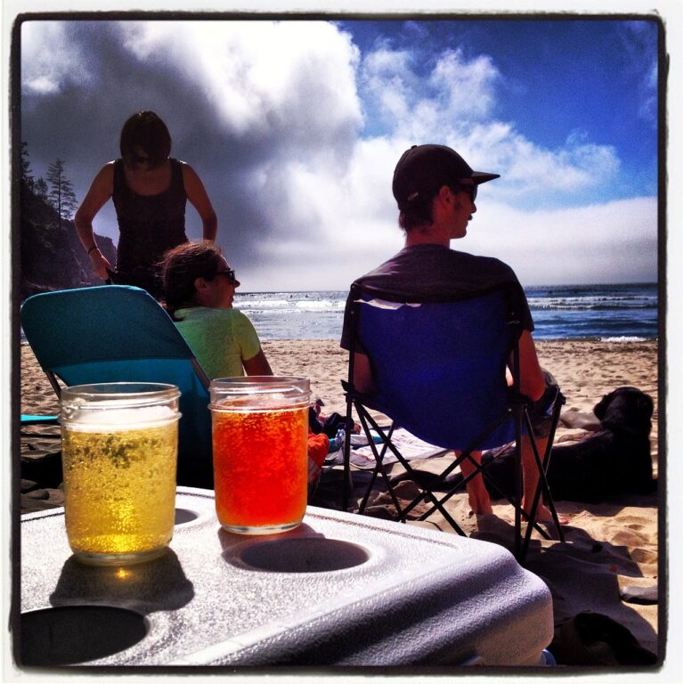 People relaxing on a sunlit beach with two drinks in the foreground and the ocean in the background.