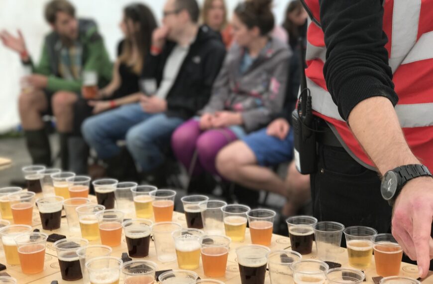 Rows of beer samples in clear cups on a table in the foreground, with a blurred group of people sitting in the background, apparently at a beer tasting event.