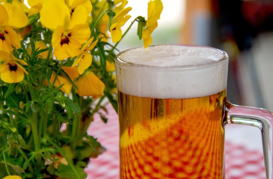 A full mug of beer with foam on top, placed in front of a bright yellow pansy flowers, on a table with a pink and white checkered tablecloth.