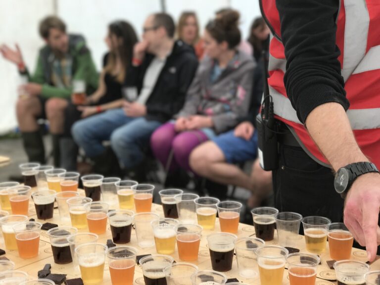 Rows of beer samples in clear plastic cups on a table in the foreground, with blurry background of people sitting and interacting at an event tent. A person in a striped shirt with a walkie-talkie is partially visible on the right.