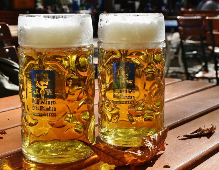 Two full steins of beer with frothy heads on a wooden table, outdoor setting with chairs in the background, showcasing Augustiner-Bräu München logos.