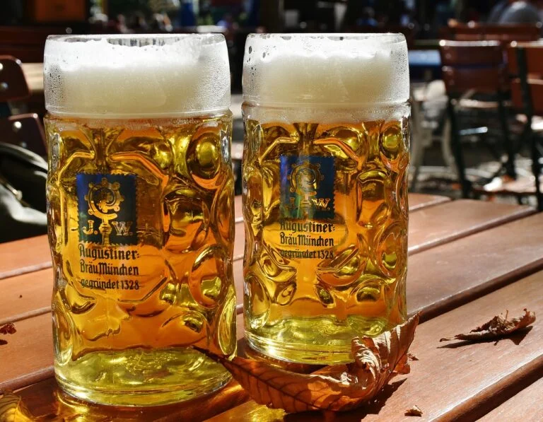 Two full steins of beer with frothy heads on a wooden table, outdoor setting with chairs in the background, showcasing Augustiner-Bräu München logos.