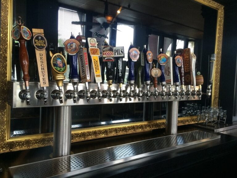 A row of assorted beer taps with various labels and handles mounted on a bar with a mirrored backdrop.