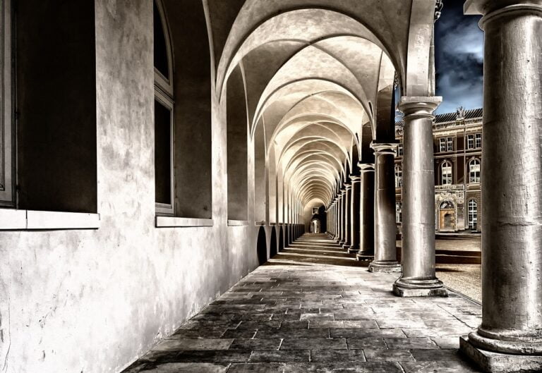 An arched corridor with columns on one side leading into the distance, characterized by strong perspective lines and contrasting light and shadow, next to a classical building.