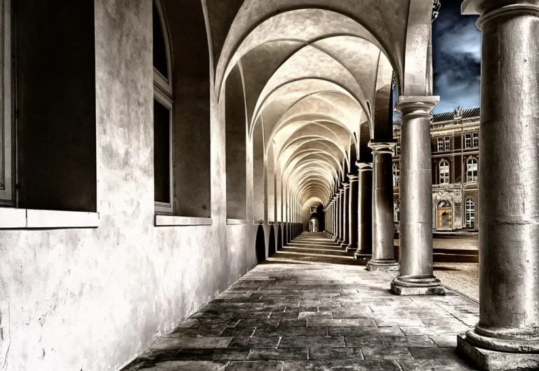An arched corridor with columns on one side leading into the distance, characterized by strong perspective lines and contrasting light and shadow, next to a classical building.