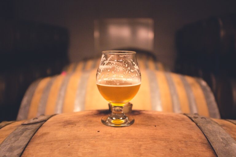 A glass of beer with foam residue on the inside sits on a wooden barrel in a dimly lit room with a blurred background of additional barrels.