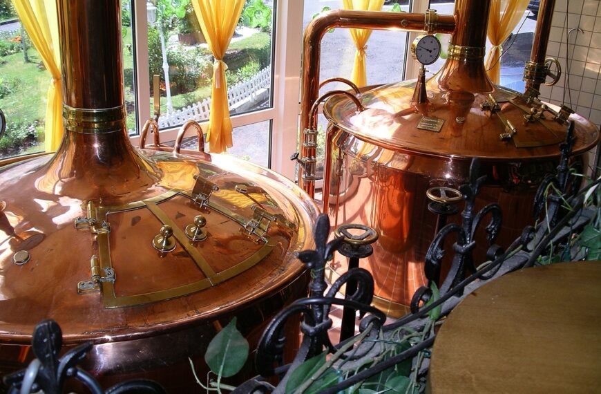 Two large copper brewing kettles with pressure gauges and pipes, situated indoors by windows with yellow curtains, surrounded by decorative metalwork and green foliage.