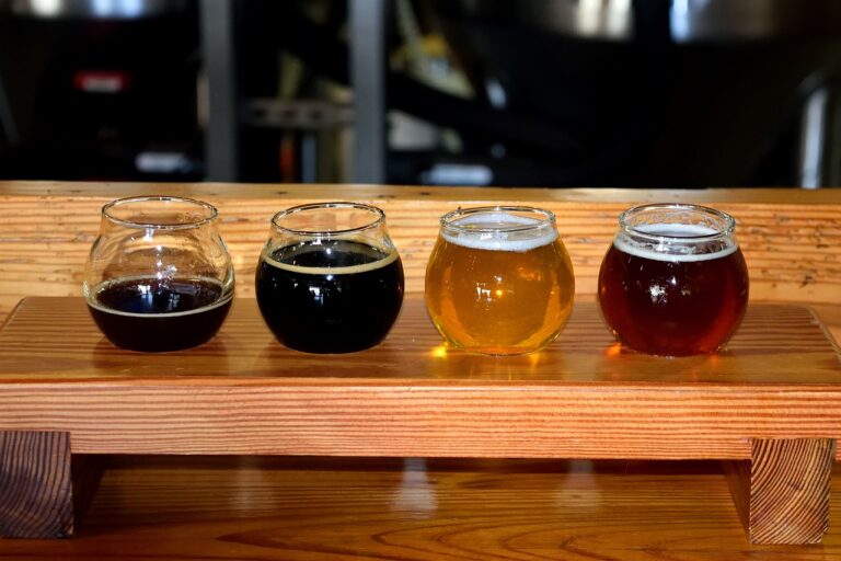 Four different types of beer in glasses arranged on a wooden flight paddle, with a brewery setting in the background.