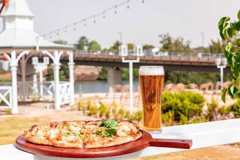A fresh pizza on a wooden board and a glass of beer on an outdoor patio table with a gazebo and string lights in the background, overlooking a bridge and river.