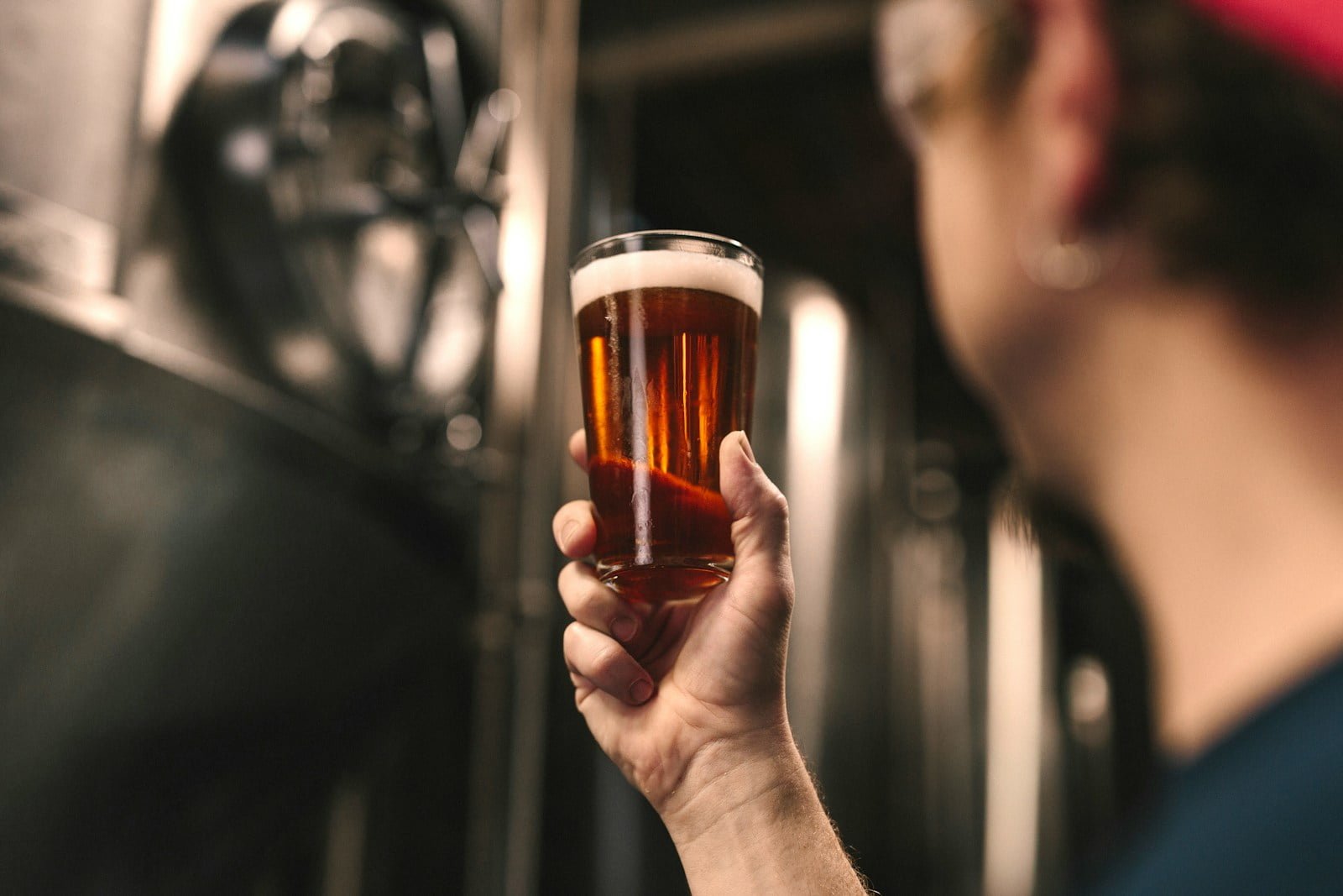 A person holding a pint glass of amber-colored beer with a brewery tank in the background.