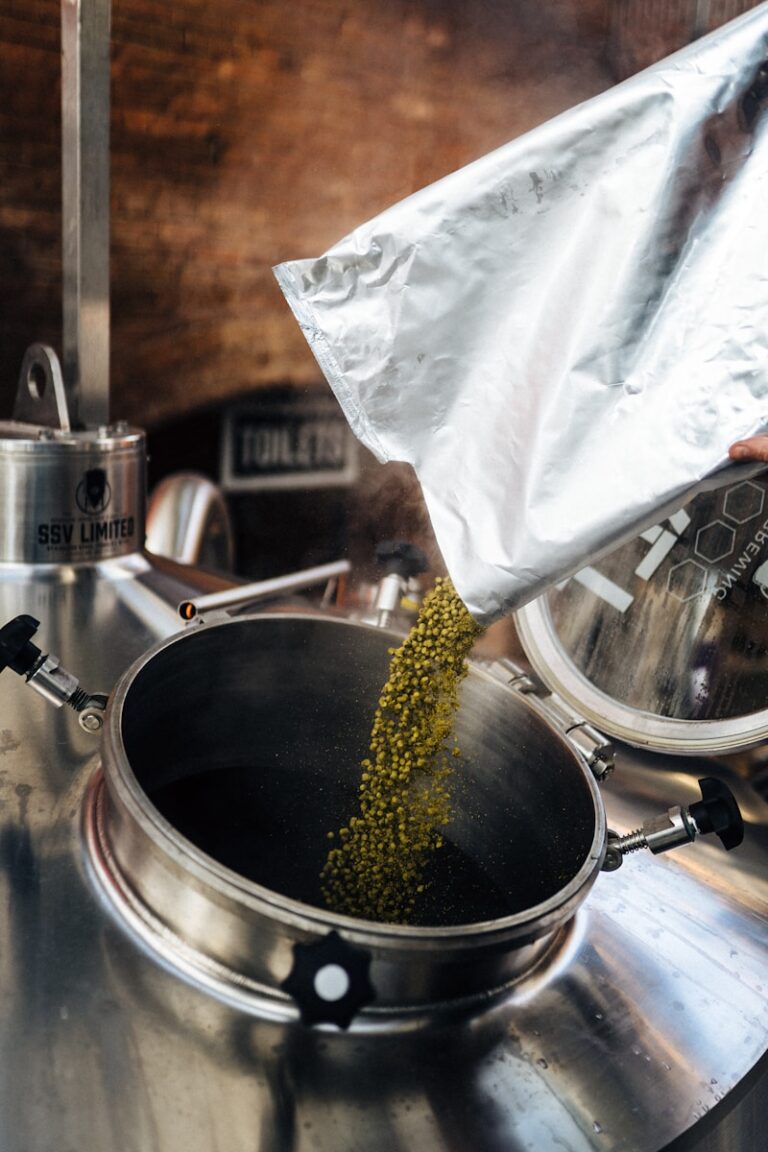 Hops being poured from a silver foil bag into a stainless steel brewing kettle in a brewery.