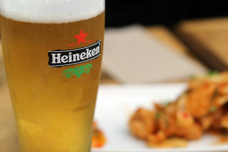 A close-up of a cold pint of Heineken beer with condensation, in focus in the foreground, and a blurry plate of fried food in the background.