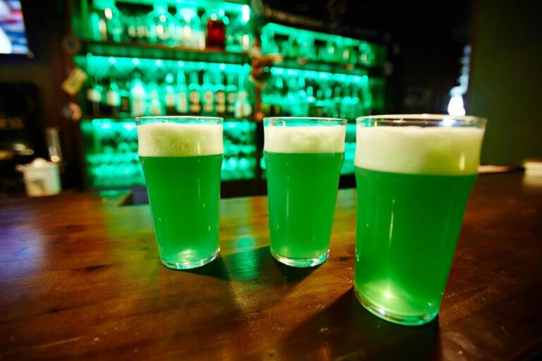 Three pints of green beer on a wooden bar top, with a backlit bar featuring green lighting in the background.