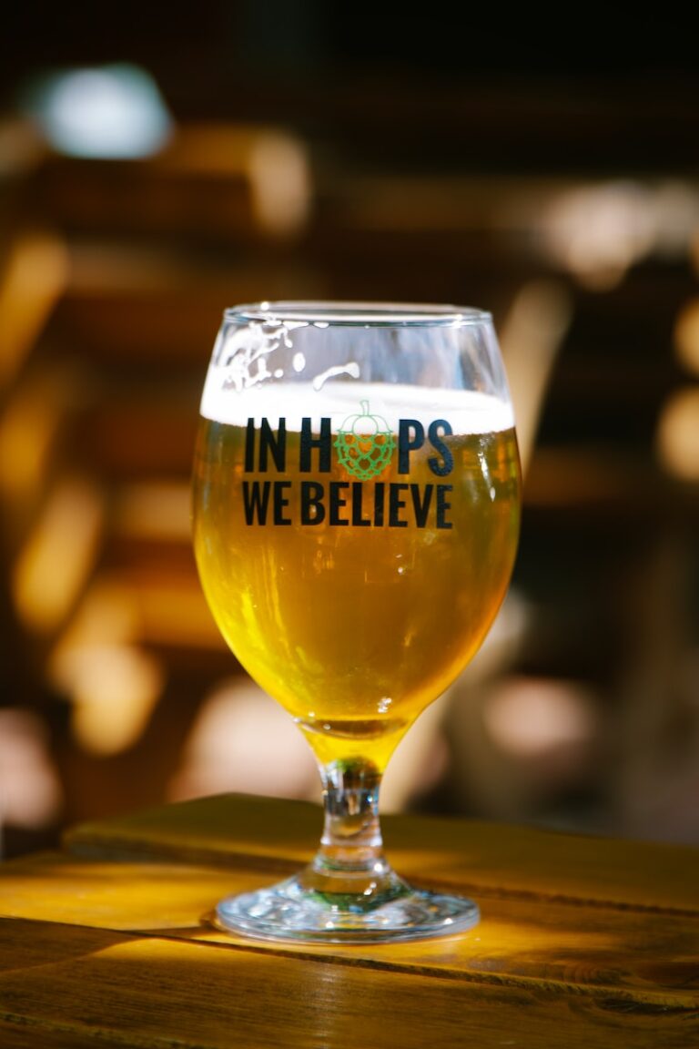 A glass of beer with the inscription "IN HOPS WE BELIEVE" on a wooden table with a blurred background.