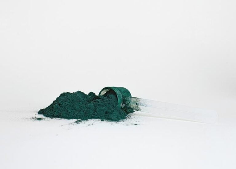 A clear scoop filled with green powder is tipped over, spilling its contents onto a white surface.