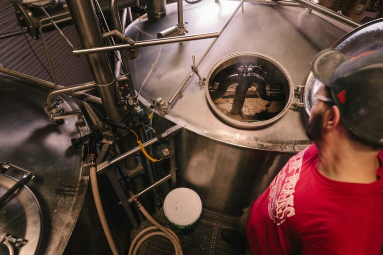 A person in a red shirt and helmet observing the inside of a large industrial brewing vat through an open hatch, with brewery equipment and pipes visible around the tank.